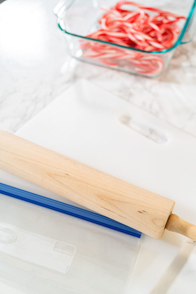 Wooden rolling pin resting on a white cutting board with a resealable plastic bag, and a glass dish filled with whole candy canes in the background.