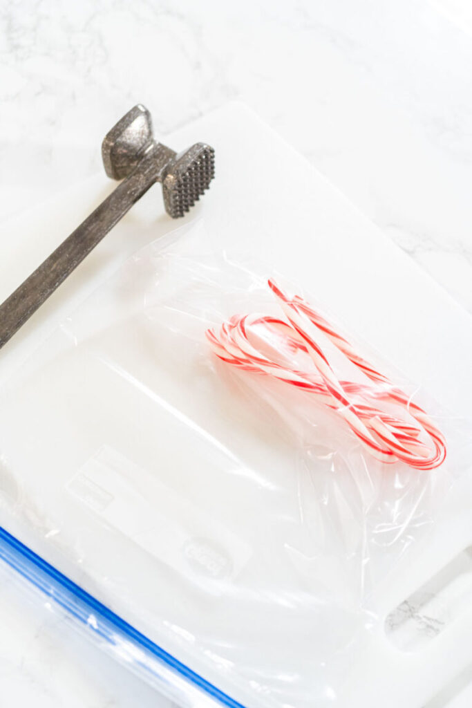 Whole candy canes inside a sealed plastic bag on a white cutting board with a metal meat mallet placed nearby.