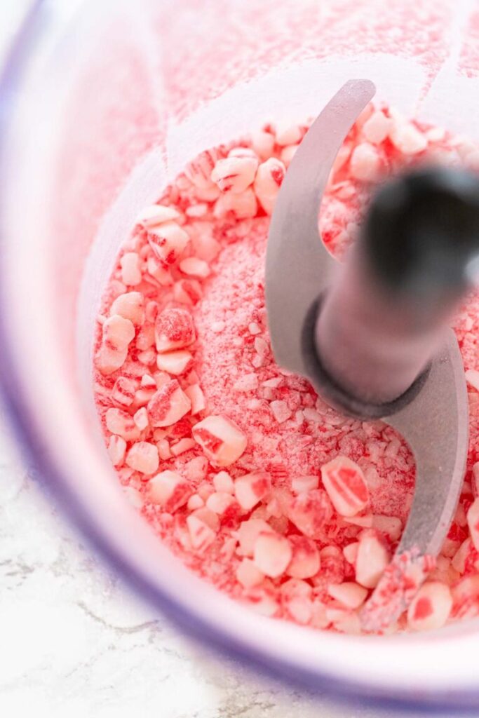 Crushed candy canes in a food processor bowl, with fine powder and small peppermint chunks ready for holiday baking.