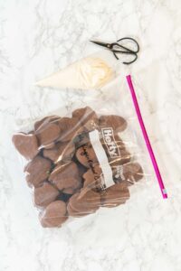 Chocolate mitten-shaped cookies stored in a large Ziploc bag, with a piping bag filled with white icing and a pair of black scissors placed on a marble countertop.