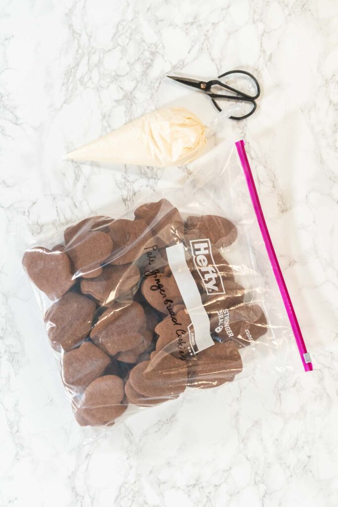 Chocolate mitten-shaped cookies stored in a large Ziploc bag, with a piping bag filled with white icing and a pair of black scissors placed on a marble countertop.