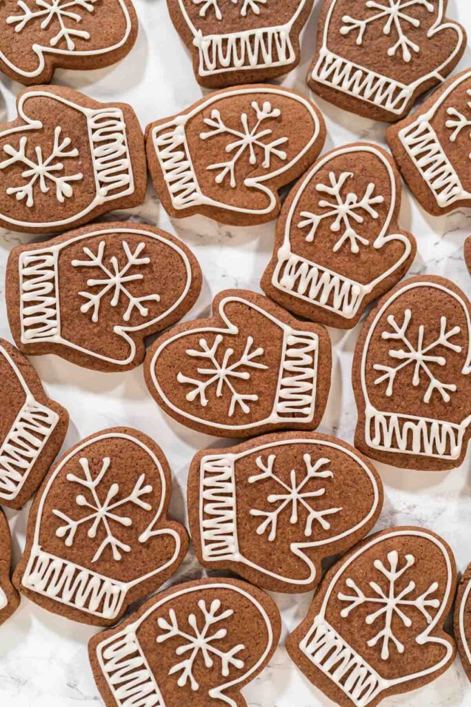 Overhead view of multiple mitten-shaped chocolate cookies decorated with white royal icing snowflakes and ribbed cuffs, arranged on a marble background.