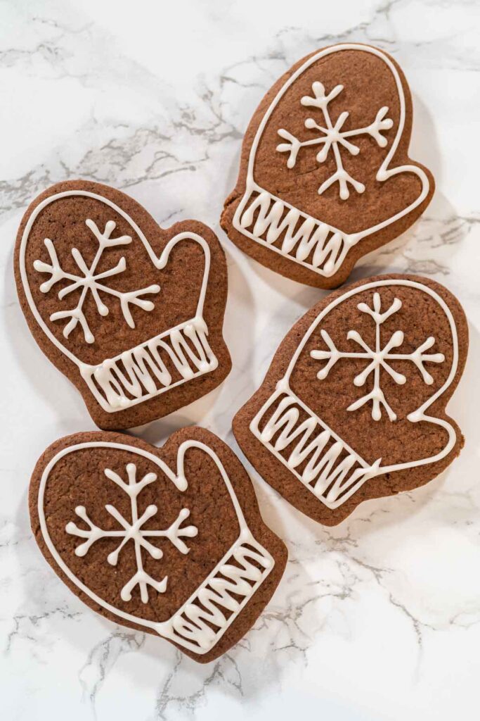 Four mitten-shaped chocolate cookies decorated with white royal icing snowflakes and cuffs, arranged on a marble surface.