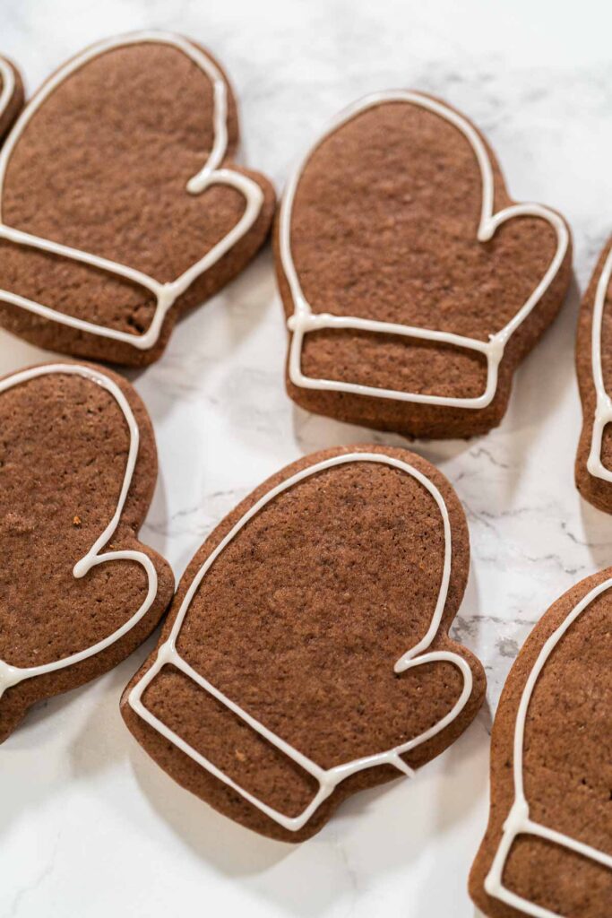 Chocolate mitten-shaped cookies outlined with white royal icing, ready for further decorating.