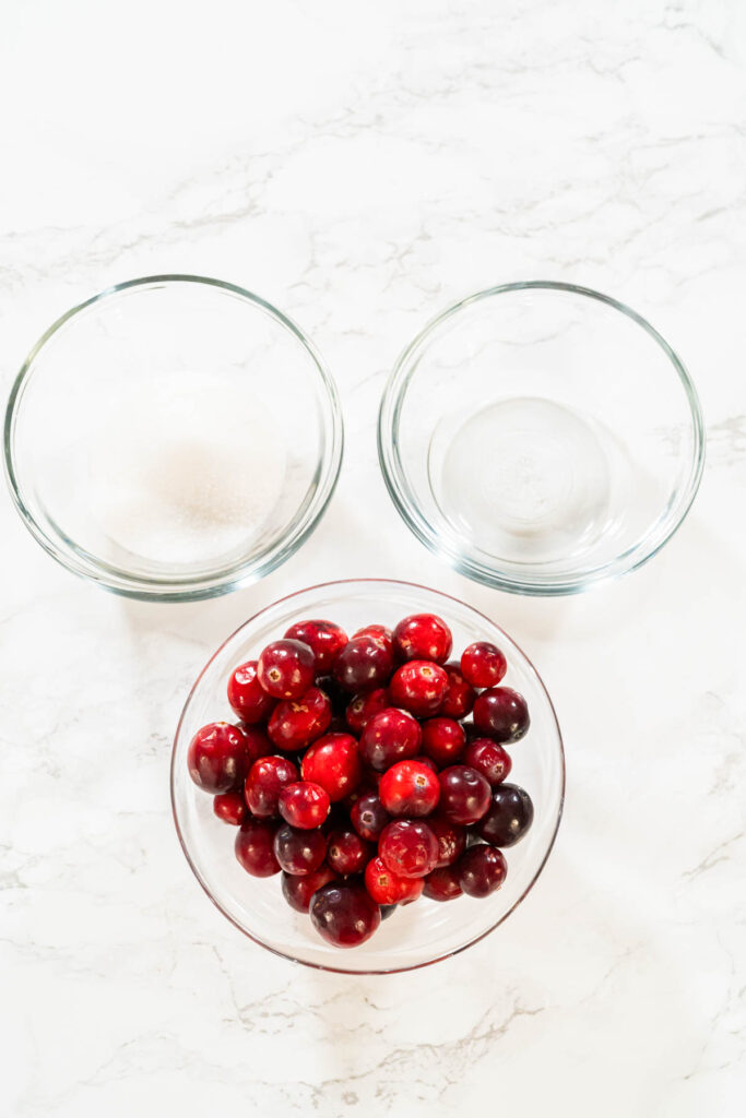 Overhead view of three glass bowls containing ingredients for cranberry puree: one with fresh whole cranberries, one with granulated sugar, and one empty bowl on a white surface.