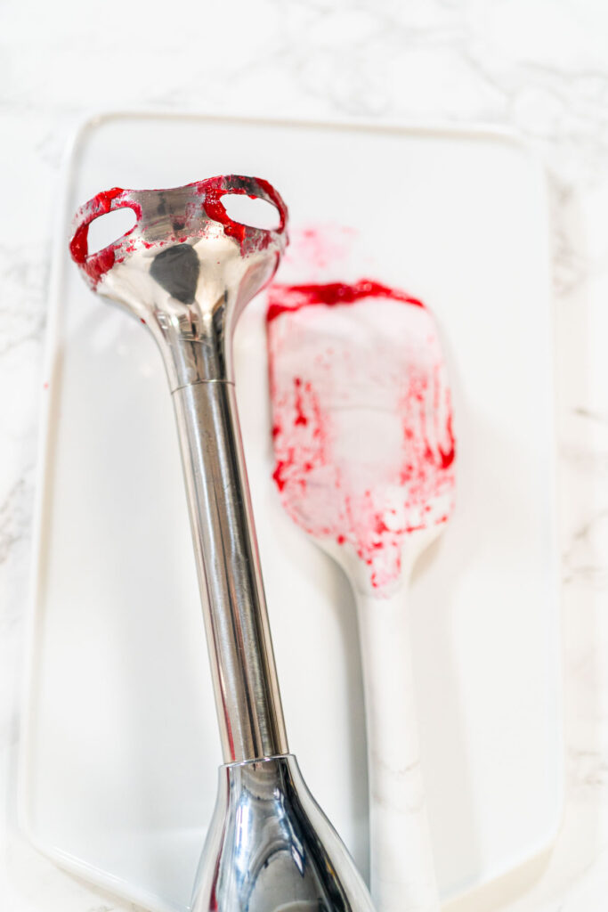 An immersion blender and silicone spatula with cranberry puree residue, showing tools used to blend and scrape the mixture for a smooth consistency.