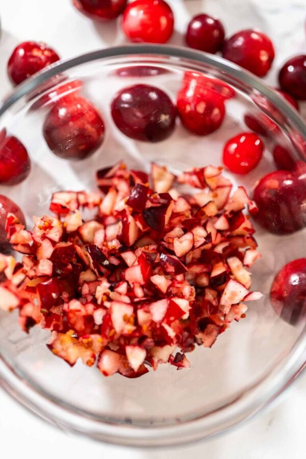Close-up of freshly diced cranberries in a glass bowl with whole cranberries scattered around, showing the vibrant red fruit pieces ready for baking or cooking.