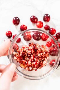 Hand holding a glass bowl filled with freshly diced cranberries, with whole cranberries scattered in the background on a white surface.