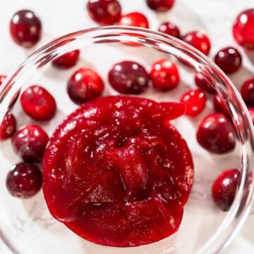 Glass bowl filled with smooth cranberry puree, surrounded by fresh whole cranberries on a white surface.