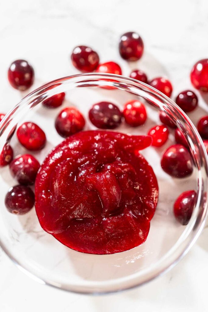Glass bowl filled with smooth cranberry puree, surrounded by fresh whole cranberries on a white surface.