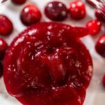 Close-up of rich, glossy cranberry puree surrounded by fresh whole cranberries, highlighting its smooth texture and vibrant red color.