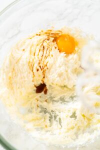 Egg yolk and vanilla extract added to a fluffy creamed mixture of butter, sugar, and cream cheese in a glass mixing bowl for lemon spritz cookie dough.