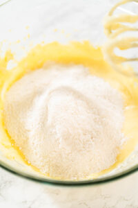 Sifted dry ingredients including flour, baking powder, salt, and cream of tartar piled on top of the lemon cookie batter in a glass mixing bowl, ready to be mixed into dough.