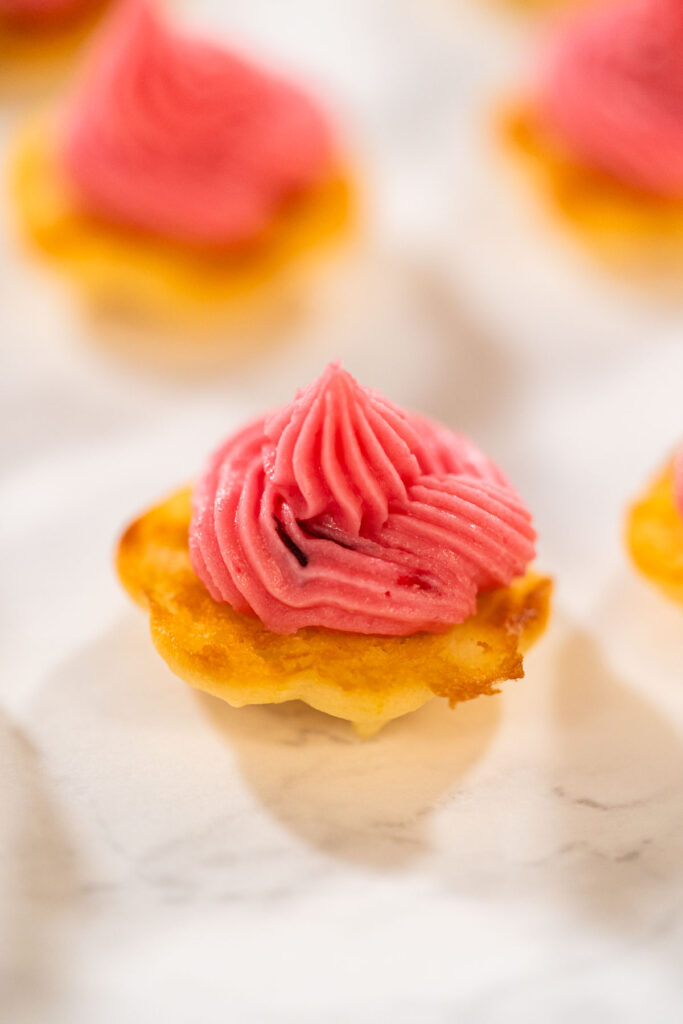 Flat cookies on the counter, with cranberry buttercream being piped onto them from a piping bag.