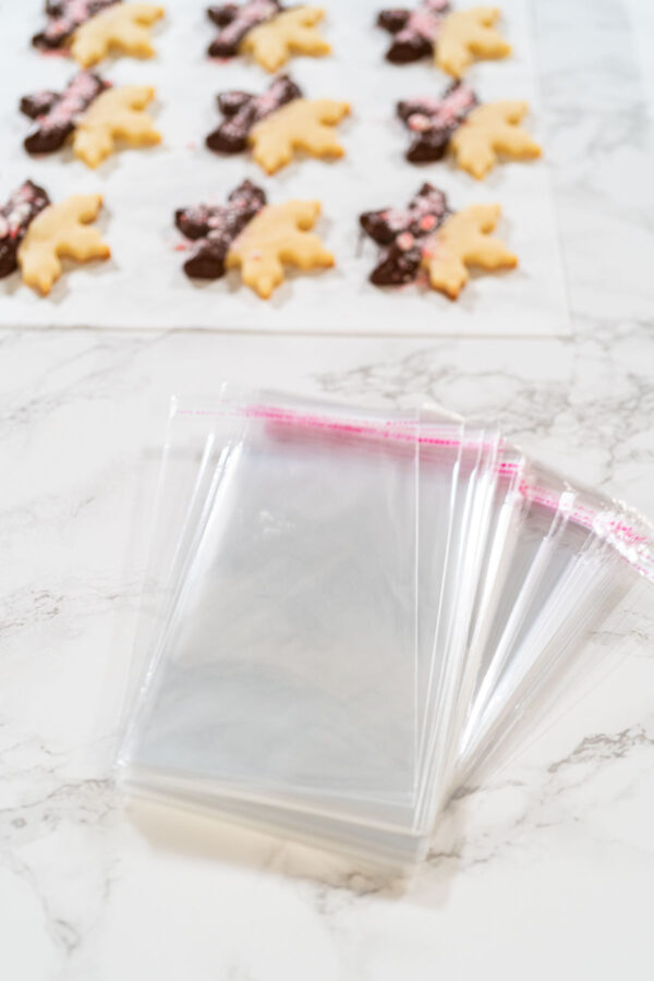 Clear treat bags stacked on a marble counter, ready for packaging peppermint chocolate-dipped snowflake cookies in the background.