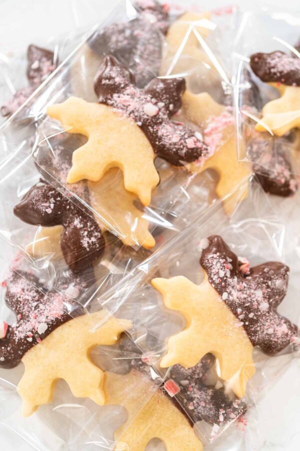 Chocolate dipped peppermint cut-out sugar cookies shaped like moose, packaged in clear treat bags for holiday gifting. Half of each cookie is coated in dark chocolate and sprinkled with crushed candy cane pieces.