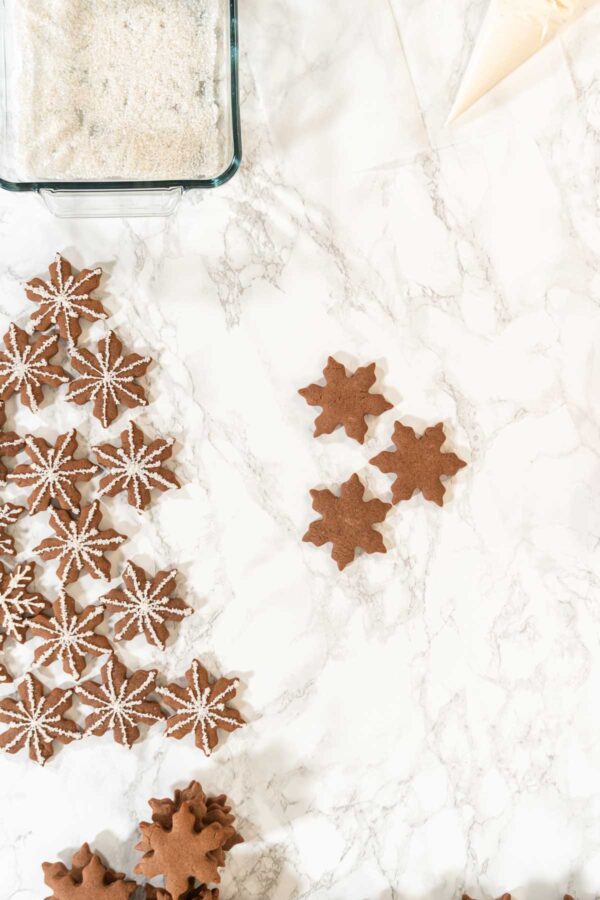 Overhead view of chocolate snowflake cookies in the process of being decorated with royal icing, featuring piped cookies on the left, plain cookies on the right, a piping bag, and a dish of white sanding sugar on a marble surface.