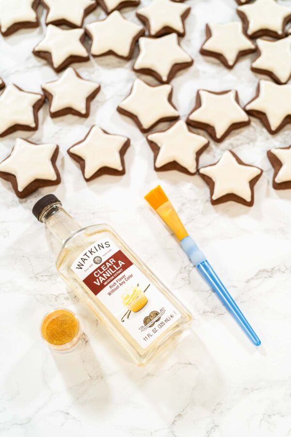 Flat lay of iced chocolate star cookies with decorating tools including Watkins clear vanilla extract, edible gold dust, and a paintbrush—perfect setup for adding shimmer to holiday cookies.