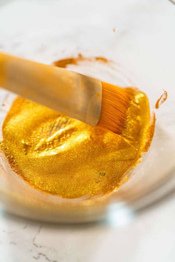 Close-up of a food-safe brush dipped in shimmering edible gold luster dust, mixed with clear vanilla extract—ready for painting cookies with a festive metallic finish.