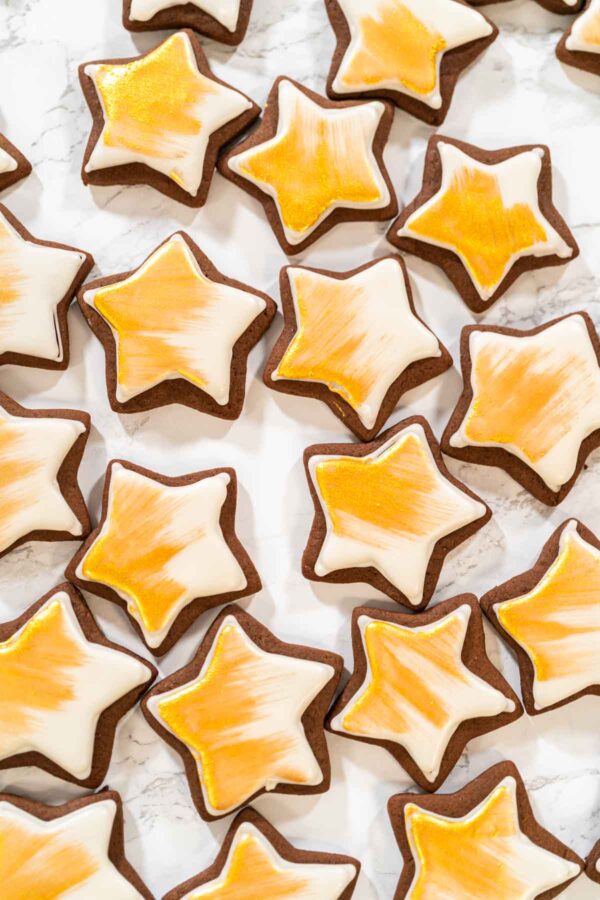 Overhead view of iced star cookies painted with edible gold luster dust, arranged on a marble surface—perfect for festive holiday cookie boxes or dessert tables.