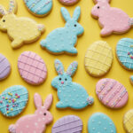 ChatGPT said: Colorful Easter sugar cookies shaped like bunnies and eggs decorated with pastel royal icing and sprinkles on a bright yellow background.