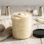 Glass jar filled with creamy Italian buttercream frosting on a light wooden surface with baking tools.