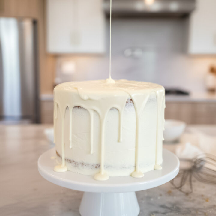 065-featured image24 - Arina Photography Smooth white chocolate ganache being poured over a frosted layer cake on a white cake stand in a bright kitchen setting.