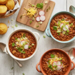 Bowls of hearty turkey chili topped with shredded cheese, radish slices, green onions, and cilantro, served with golden cornbread muffins on a rustic white table.